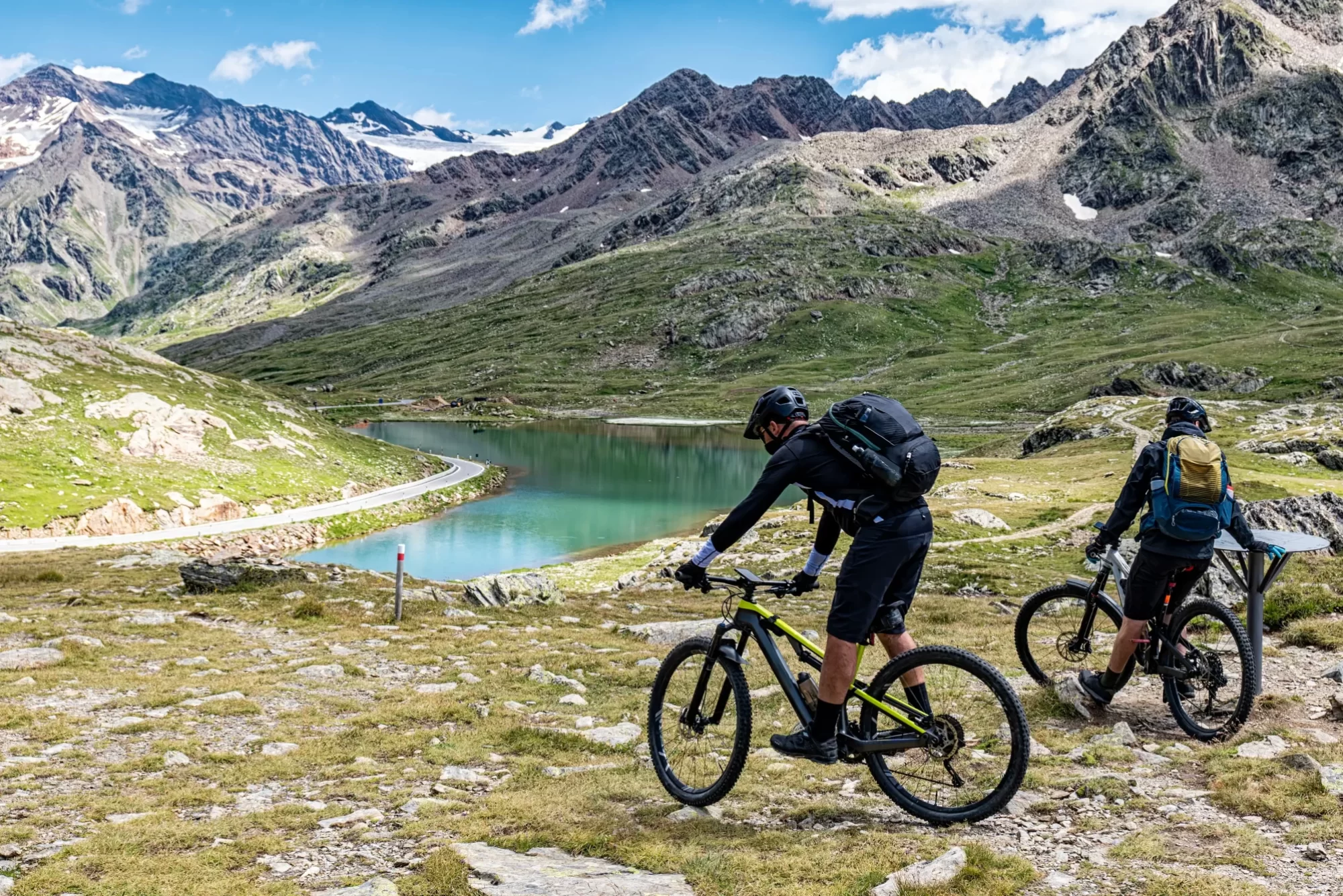 Ciclisti su mountain bike esplorano un paesaggio alpino con un lago e montagne sullo sfondo.