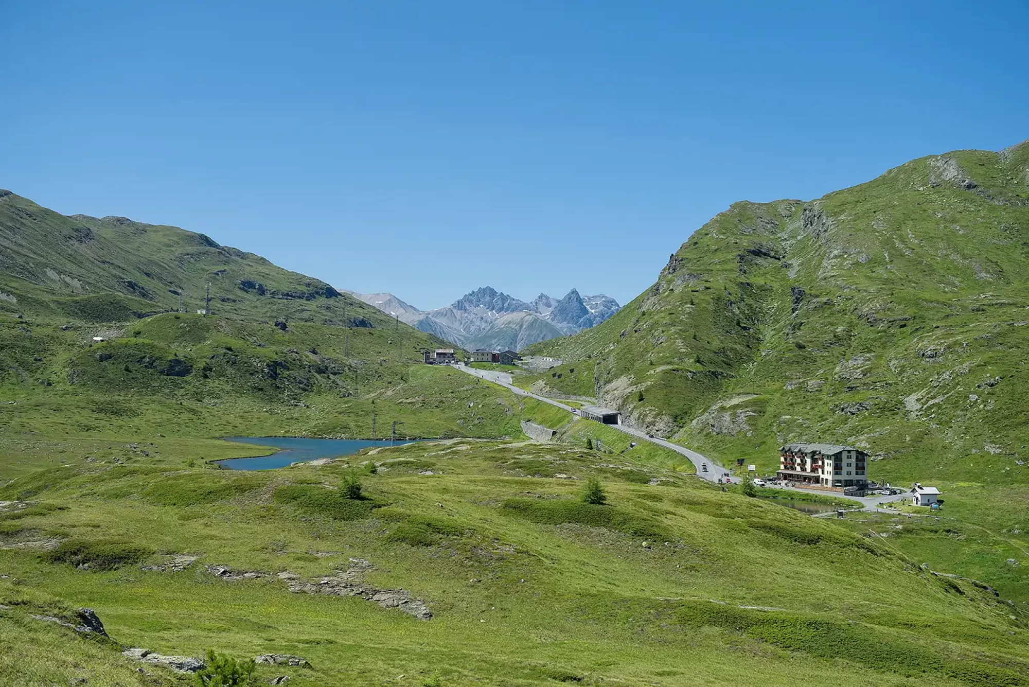 Vista panoramica di un paesaggio montano con una strada, un piccolo lago e verdi colline.