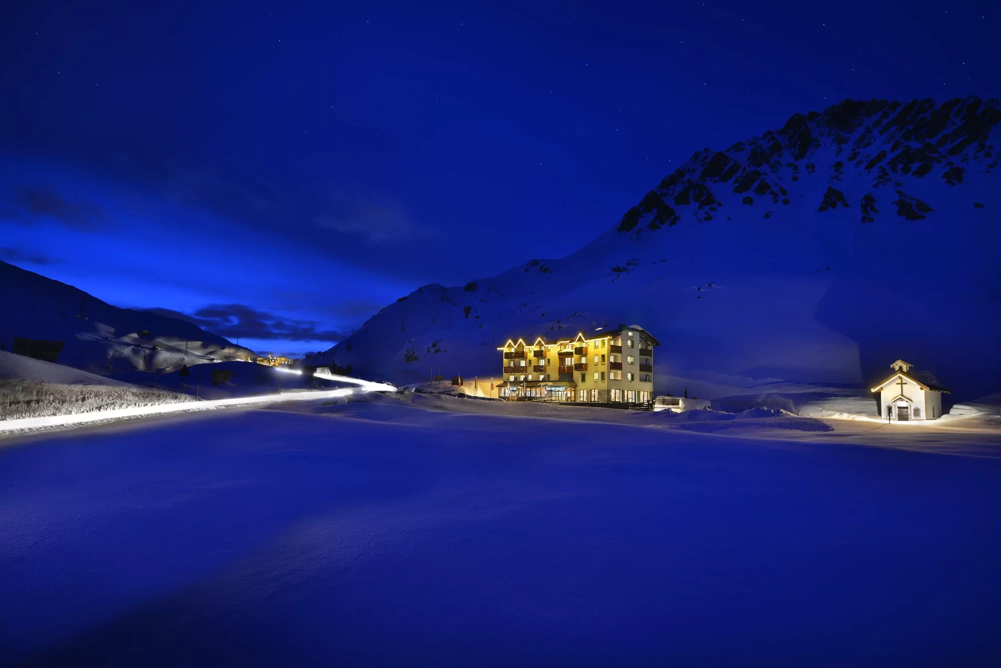 Vista notturna di un hotel illuminato e una cappella in una montagna innevata sotto un cielo blu.