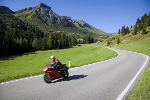 Un motociclista in giacca rossa percorre una strada di montagna in una giornata soleggiata.