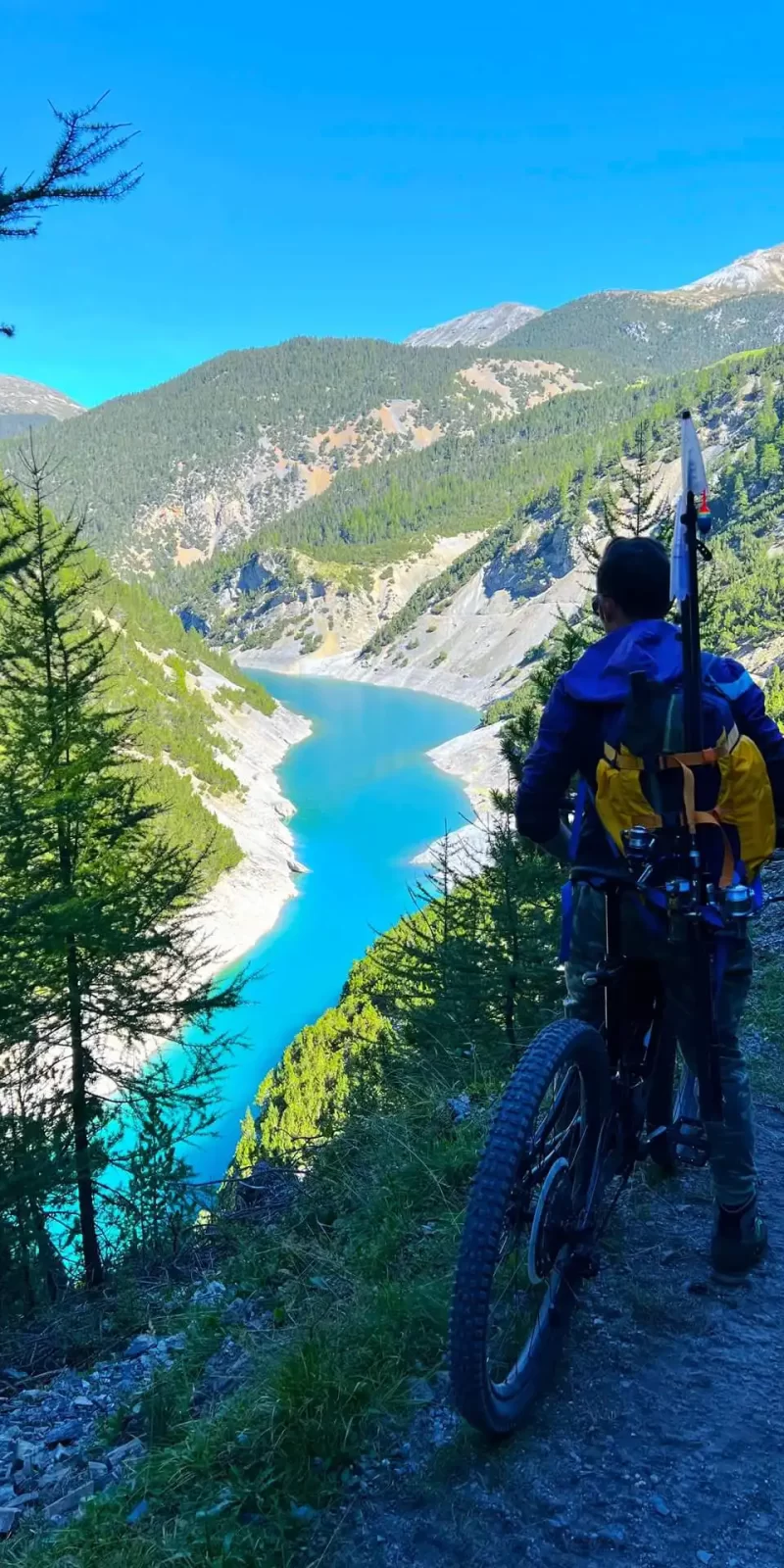 Persona in mountain bike su un sentiero panoramico con vista su un lago alpino e montagne verdi.