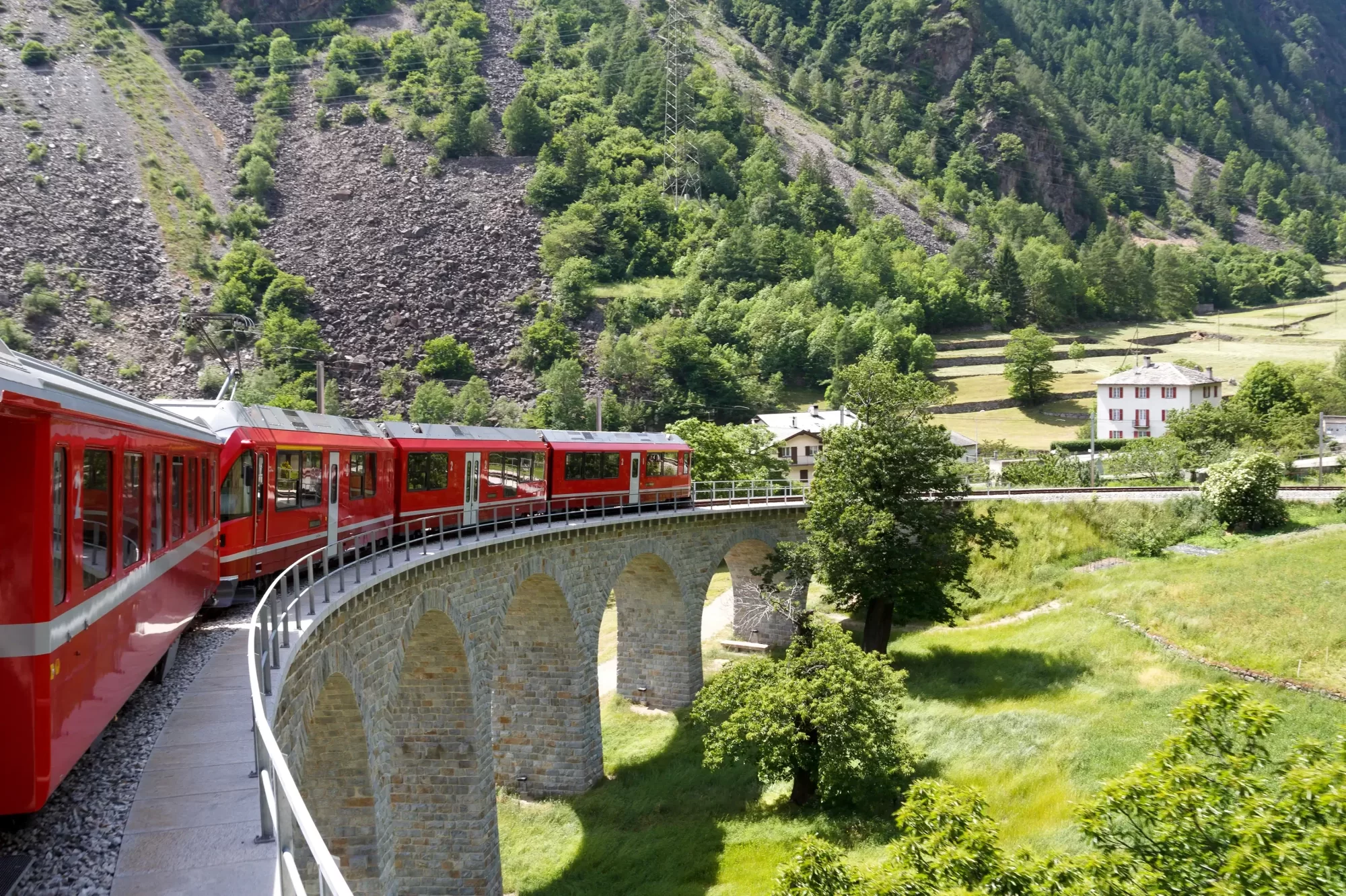 Treno rosso attraversa un viadotto in un paesaggio verde di montagna.