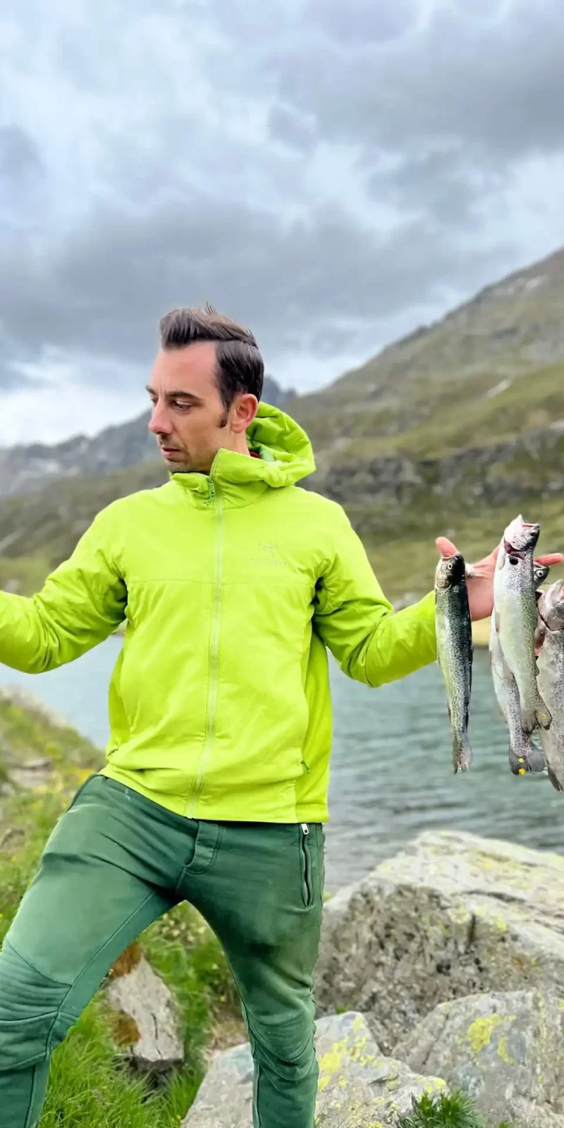 Uomo in abbigliamento verde che tiene diversi pesci in riva a un lago montano.