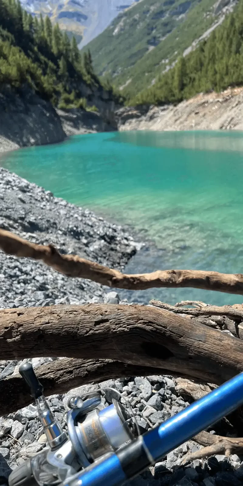 Una canna da pesca appoggiata su rami secchi vicino a un lago alpino dalle acque turchesi.