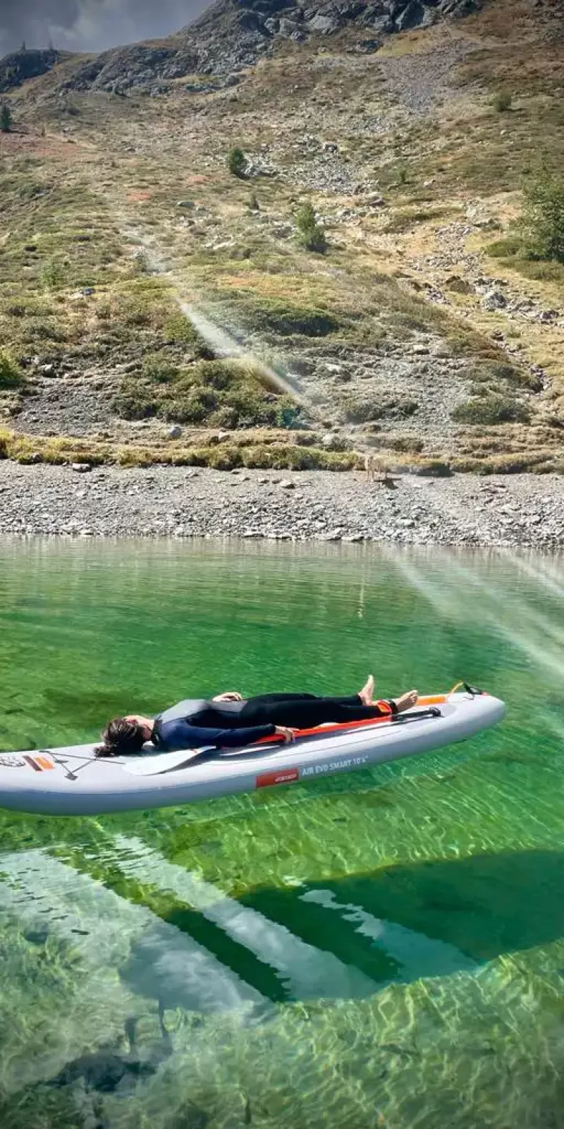 Persona distesa su una tavola da paddle in un lago limpido circondato da montagne erbose.