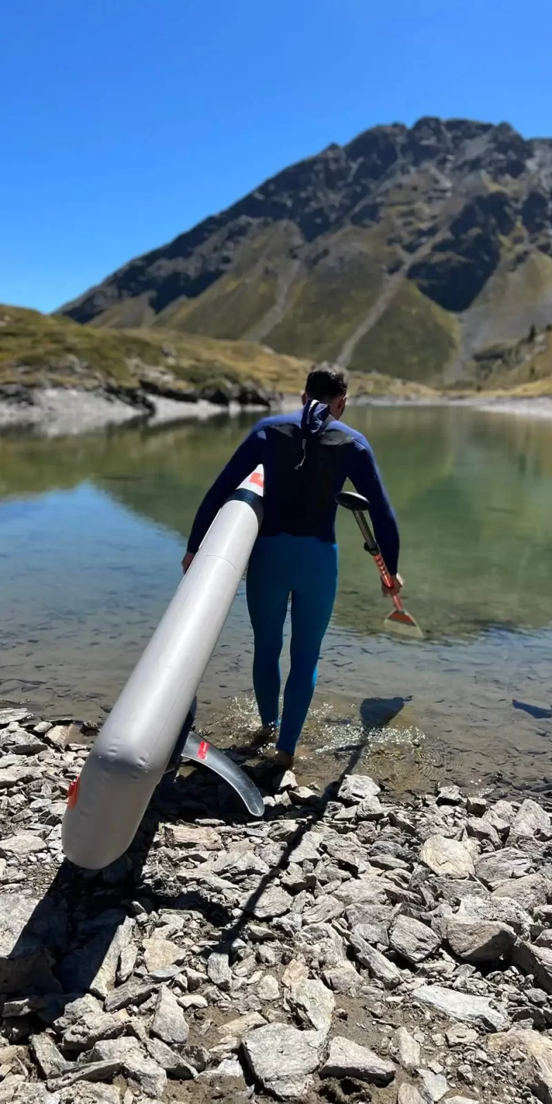 Persona porta una tavola da paddle verso un lago montano limpido.