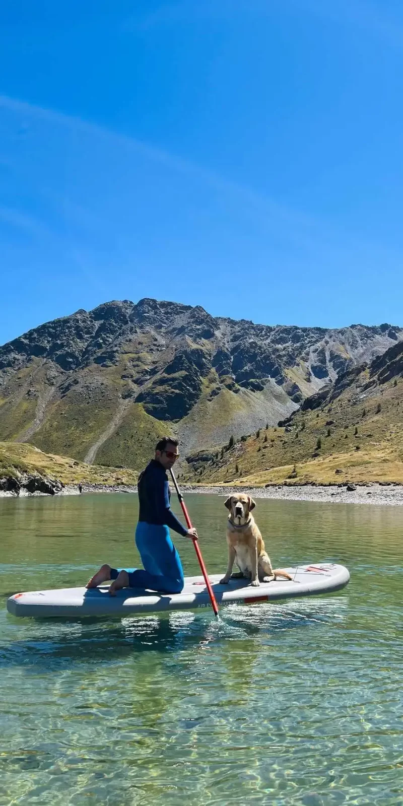 Uomo e cane su una tavola da paddle in un lago montano circondati da montagne.
