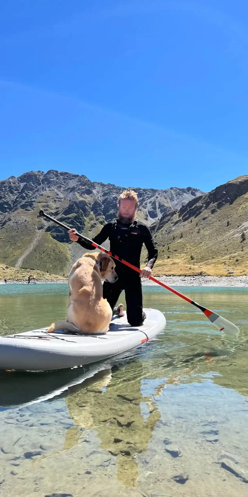 Persona e cane su un paddleboard in un lago alpino sotto un cielo blu.