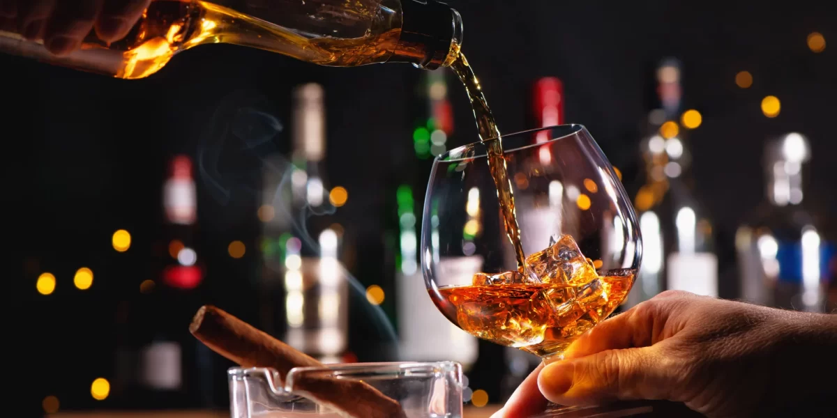 Versamento di whisky in un bicchiere con ghiaccio accanto a un posacenere con sigaro.